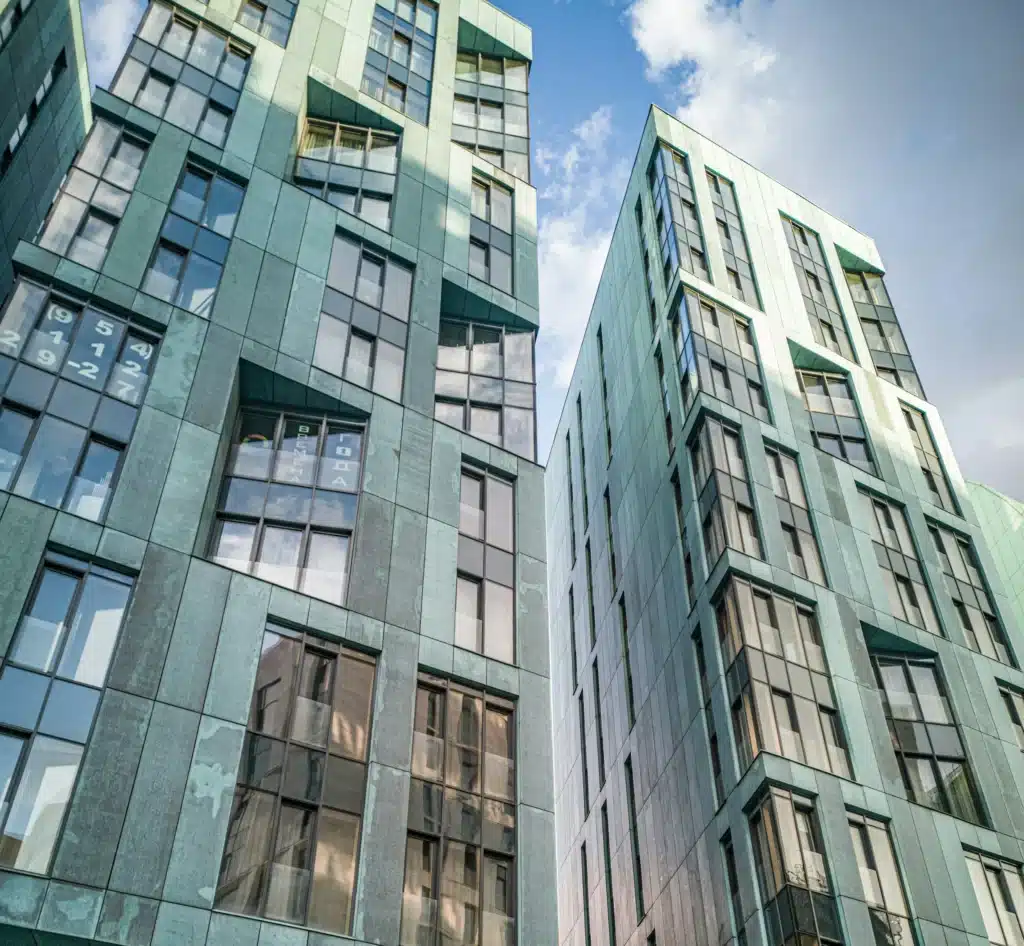 Two modern buildings with green facades and reflective windows against a blue sky with clouds.