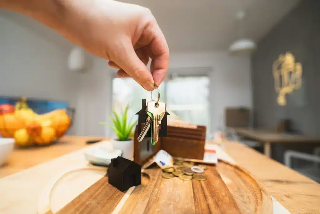 Hand holding a set of house keys with a miniature house keychain and coins scattered on a wooden surface.